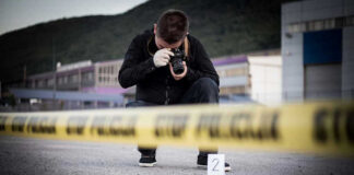 Crime scene photographer behind police tape with evidence marker.