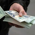 Newsom Defies Trump—$90M Giveaway A hand holding a stack of hundred dollar bills with one bill prominently displayed