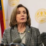 Nancy Pelosi speaking at a press conference with an American flag in the background