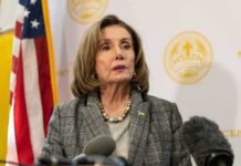 Nancy Pelosi speaking at a press conference with an American flag in the background