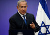 Man in suit giving speech in front of Israeli flag.