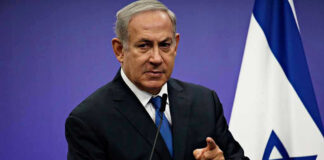 Man in suit giving speech in front of Israeli flag.