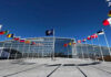 Flags outside NATO headquarters building under clear blue sky.