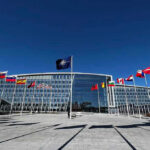 Finland Ends Nuclear Weapons Ban Flags outside NATO headquarters building under clear blue sky.