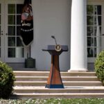 Blanche Defends Trump Pardons as Scarborough Fires Back Podium with presidential seal in a garden colonnade