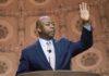Trump Declares Tucker Carlson “Irrelevant” Speaker in suit addressing audience at podium with hand raised