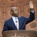 Speaker in suit addressing audience at podium with hand raised