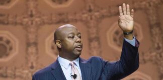Speaker in suit addressing audience at podium with hand raised