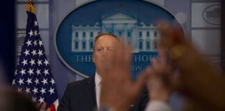 Press secretary at podium with raised hands in briefing room