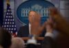 Press secretary at podium with raised hands in briefing room
