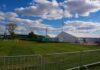 Temporary white tents and equipment on a grassy field with traffic cones and metal barriers