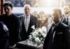 Mourners in formal attire carry a flower-adorned casket indoors