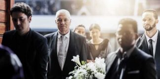 Mourners in formal attire carry a flower-adorned casket indoors