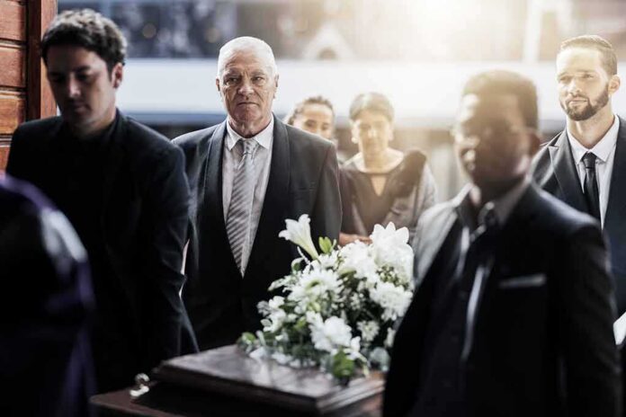 shutterstock_2224696553 Mourners in formal attire carry a flower-adorned casket indoors