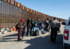 Group of people standing by a border fence with officers and vehicles