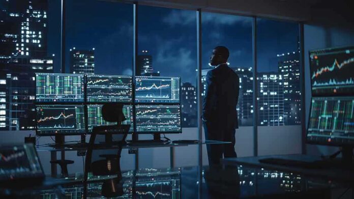 shutterstock_2088641509 Trader in a dark office with multiple stock monitors and city skyline at night